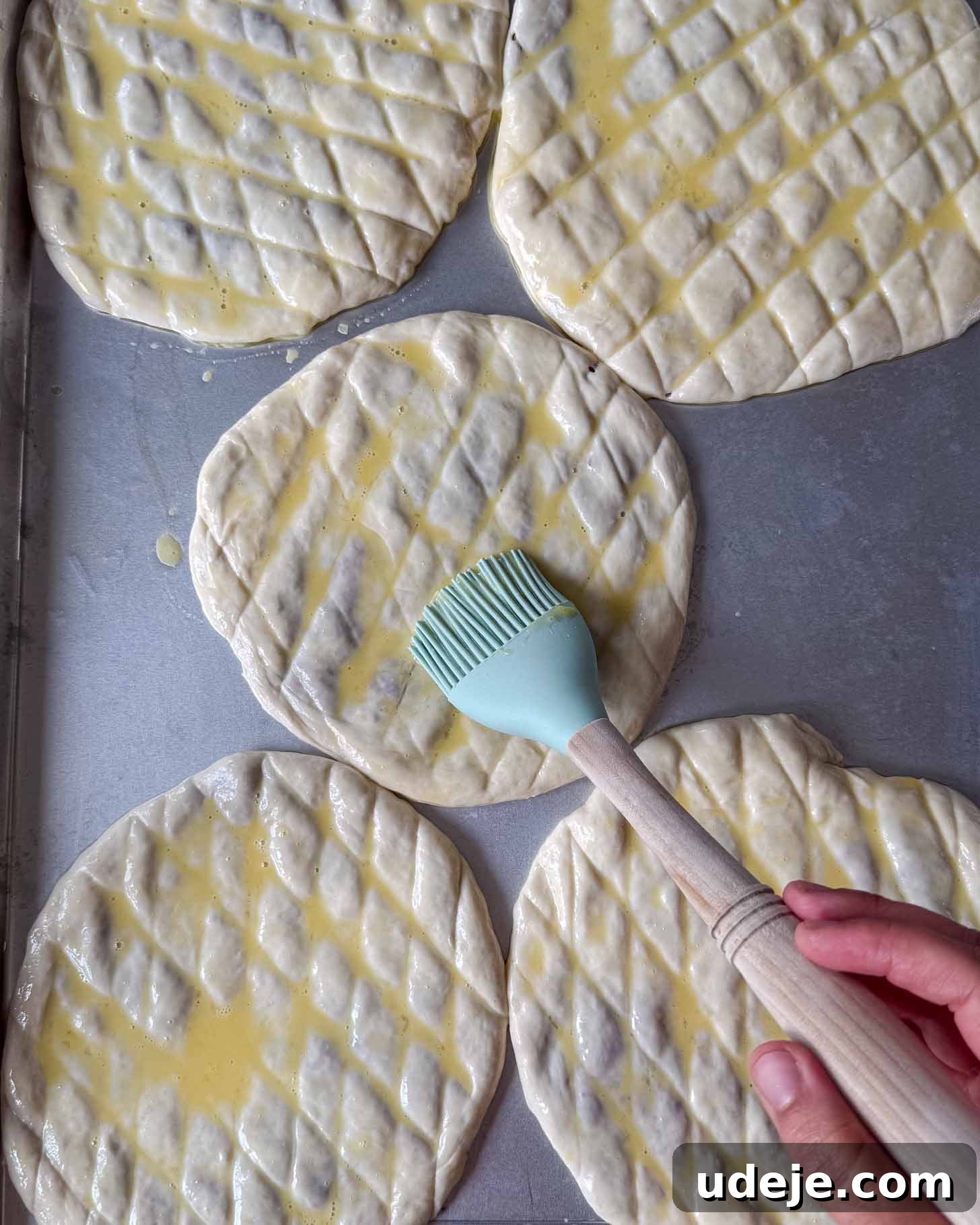 Five round pieces of dough with a diamond pattern are brushed with egg wash by a hand holding a silicone pastry brush on a metal baking sheet.