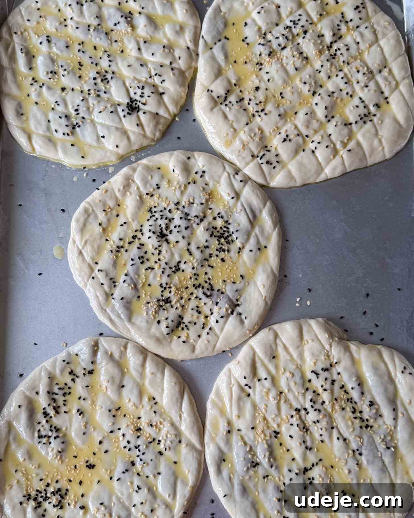 Five round, uncooked flatbreads with a crosshatch pattern, brushed with egg wash and sprinkled with black and white sesame seeds, are arranged on a metal baking tray.