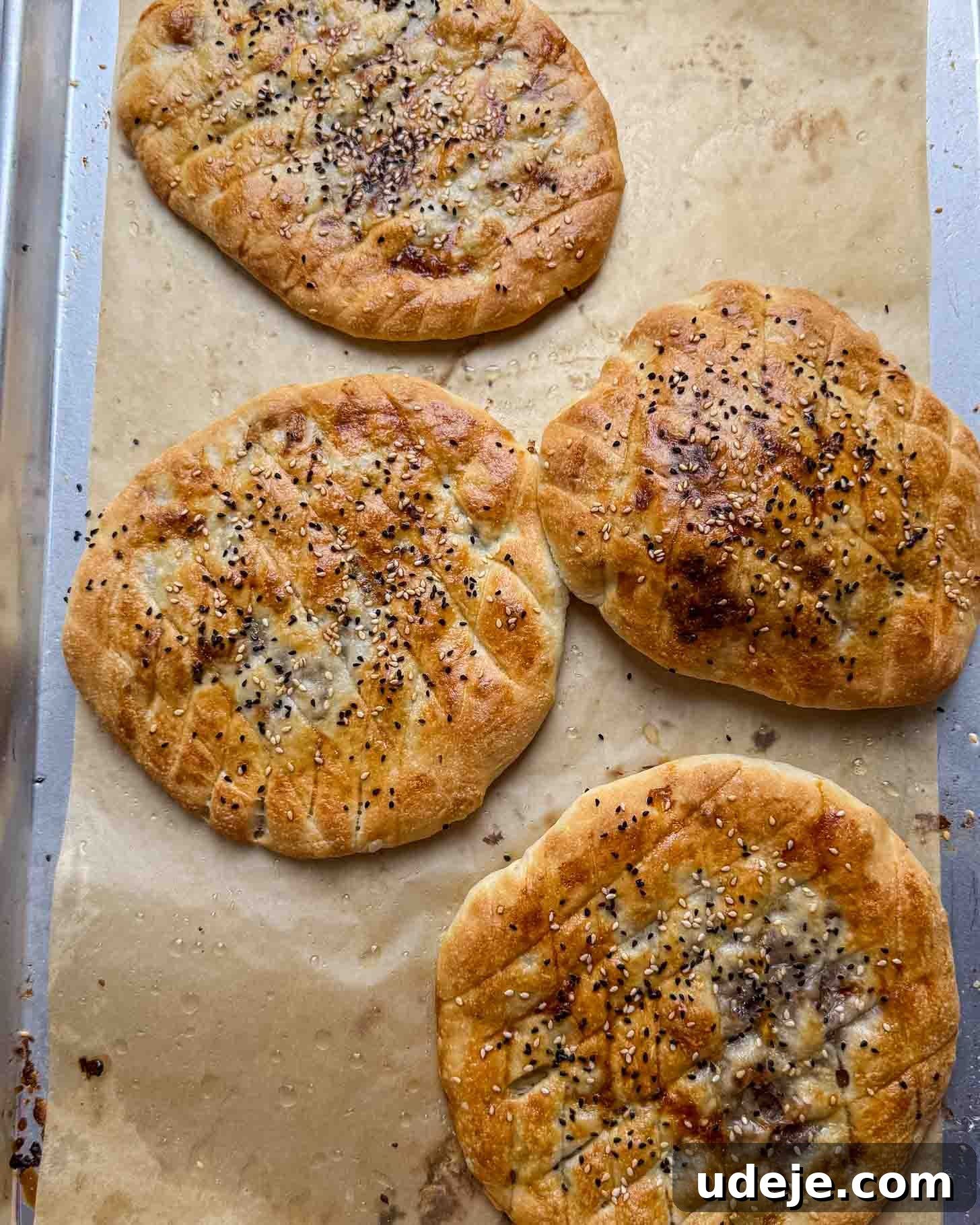 Four round, golden brown breads with sesame and black seeds on top, arranged on parchment paper on a baking tray. The breads have a crisscross pattern on the surface.
