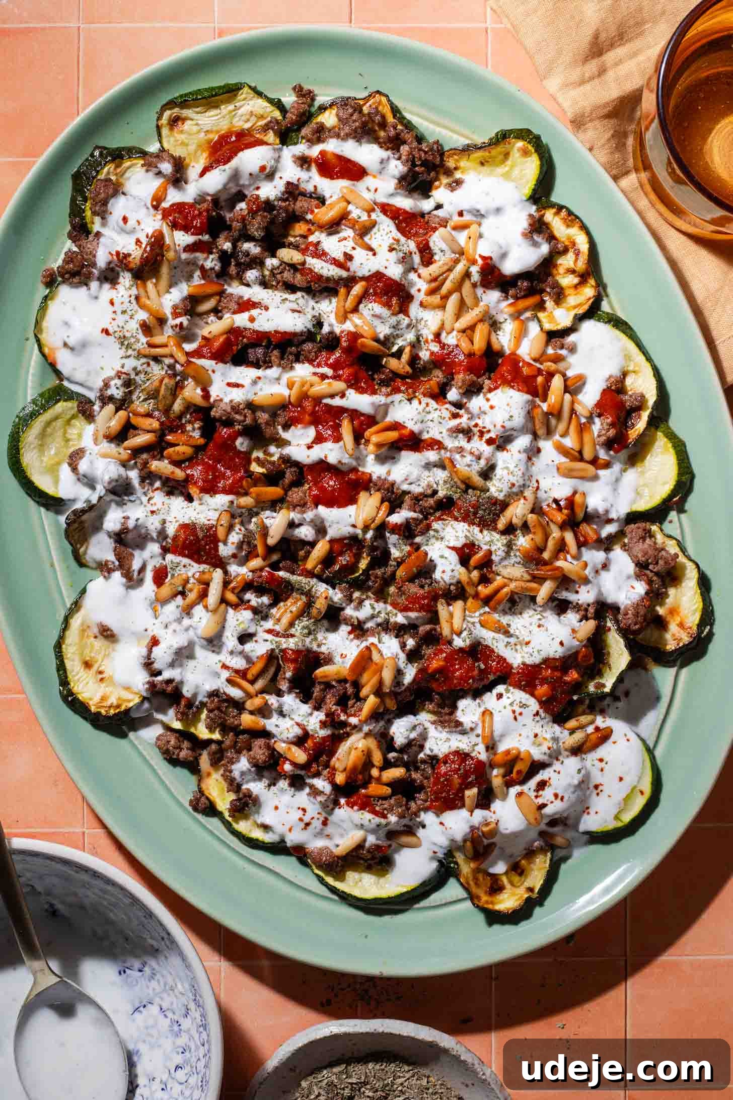 A platter of roasted zucchini slices topped with ground meat, tomato sauce, pine nuts, and a drizzle of white yogurt sauce, set on a peach-colored tiled surface.