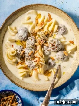 Beige speckled bowl with a creamy off white liquid, small brown meatballs covered in the off white liquid, spiral shaped white pasta and golden toasted pine nuts on top.