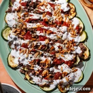 A platter of roasted zucchini slices topped with ground meat, tomato sauce, pine nuts, and a drizzle of white yogurt sauce, set on a peach-colored tiled surface.