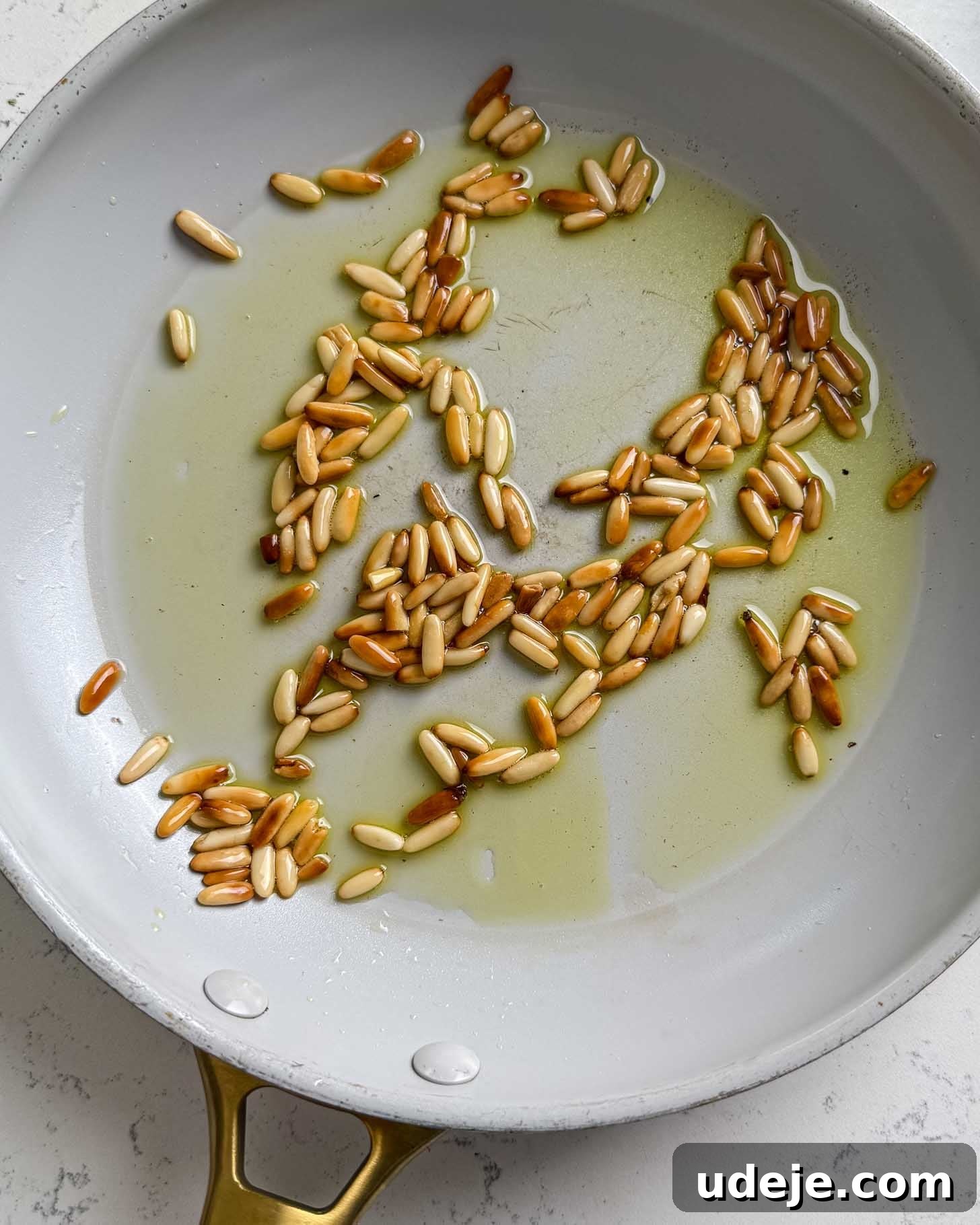 Pine nuts toasting in olive oil in a white skillet with a gold handle, some nuts turning golden brown.