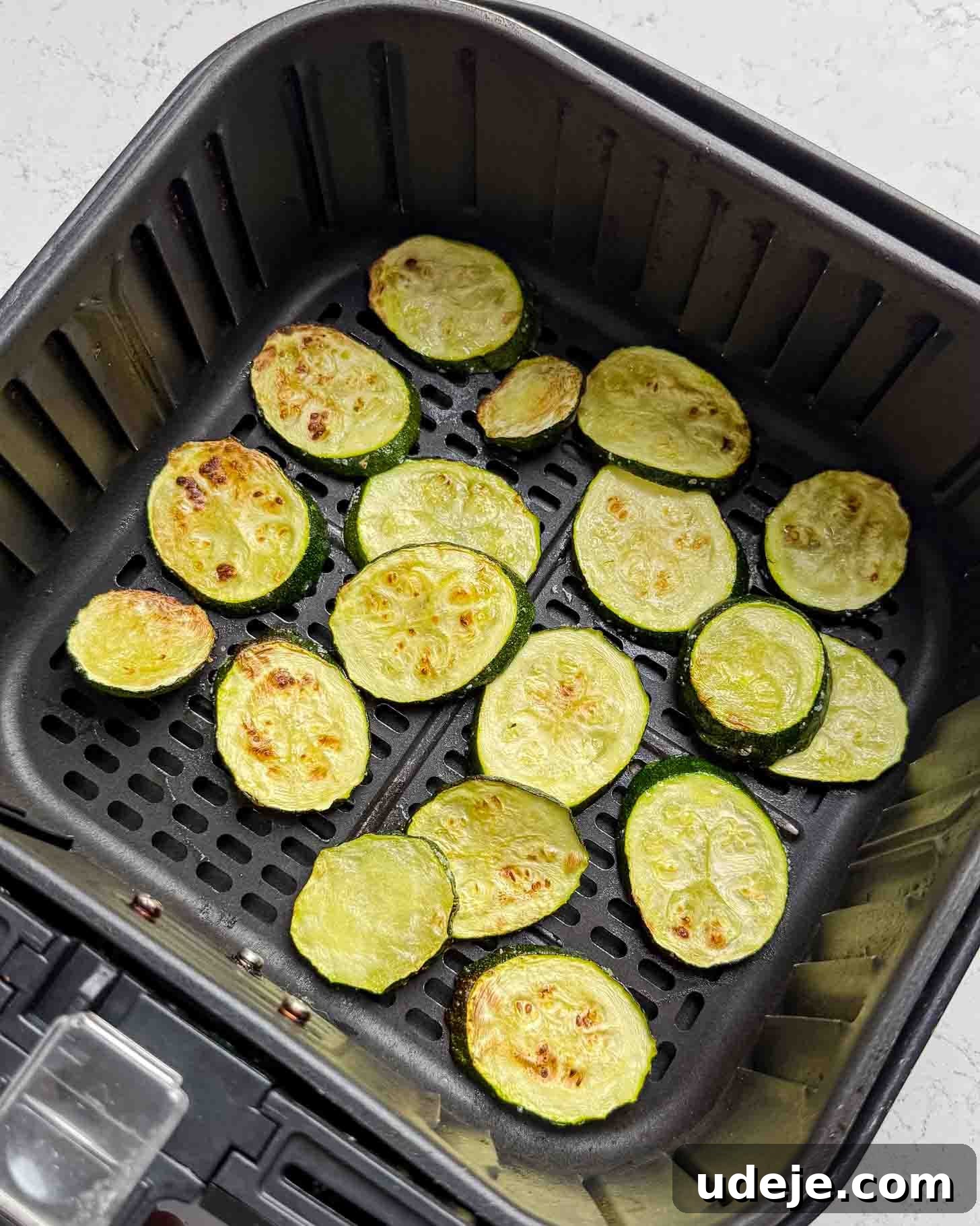 Sliced zucchini rounds, lightly browned, are arranged in a single layer inside an air fryer basket on a white countertop.