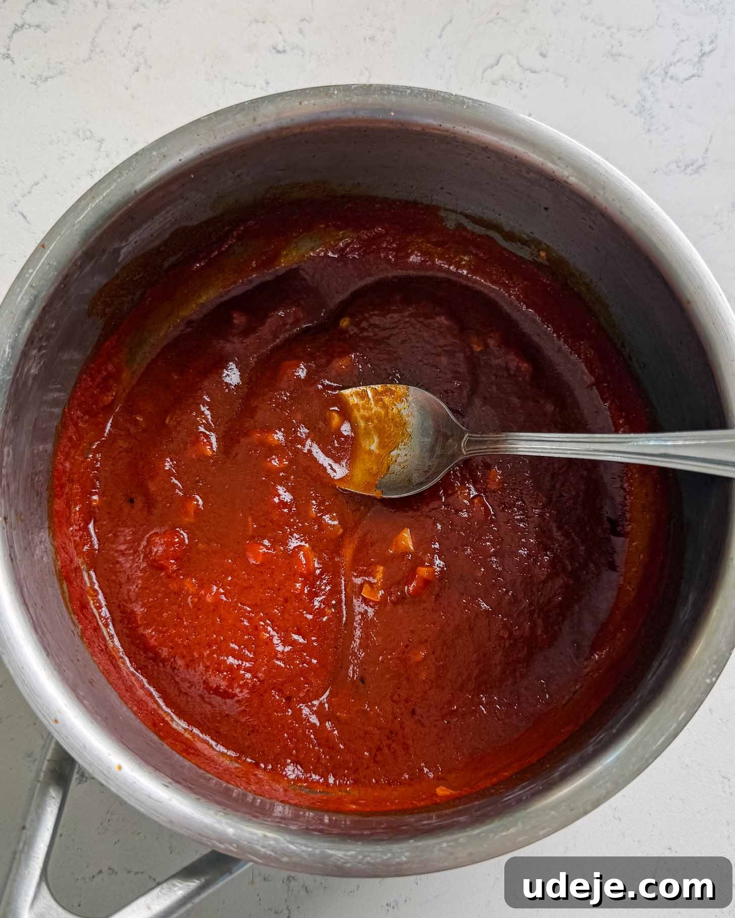 A pot of thick red barbecue sauce with a silver spoon resting inside, placed on a white countertop.