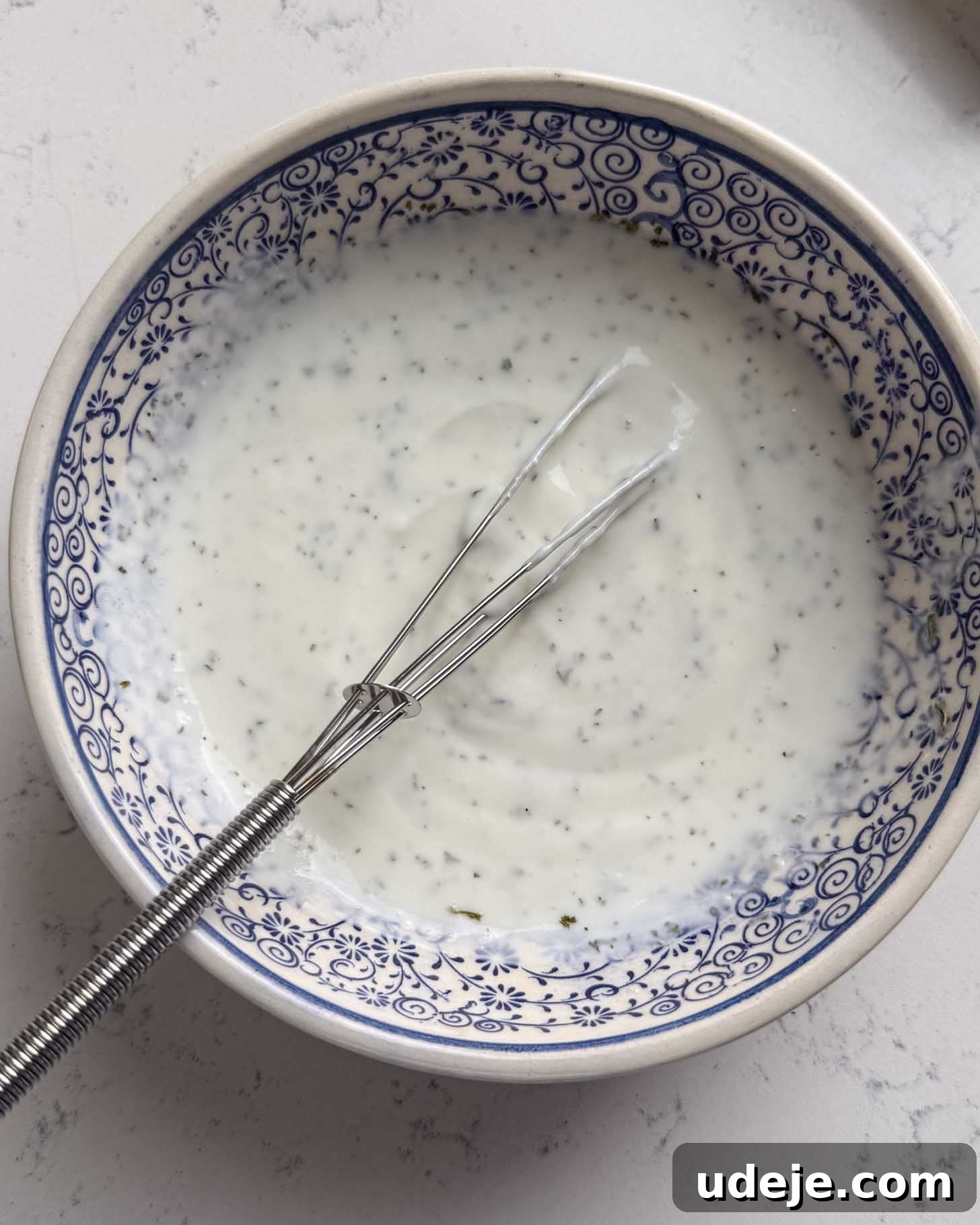 A bowl with blue patterns filled with creamy white sauce or dressing, with visible herbs, and a small metal whisk resting inside the bowl.
