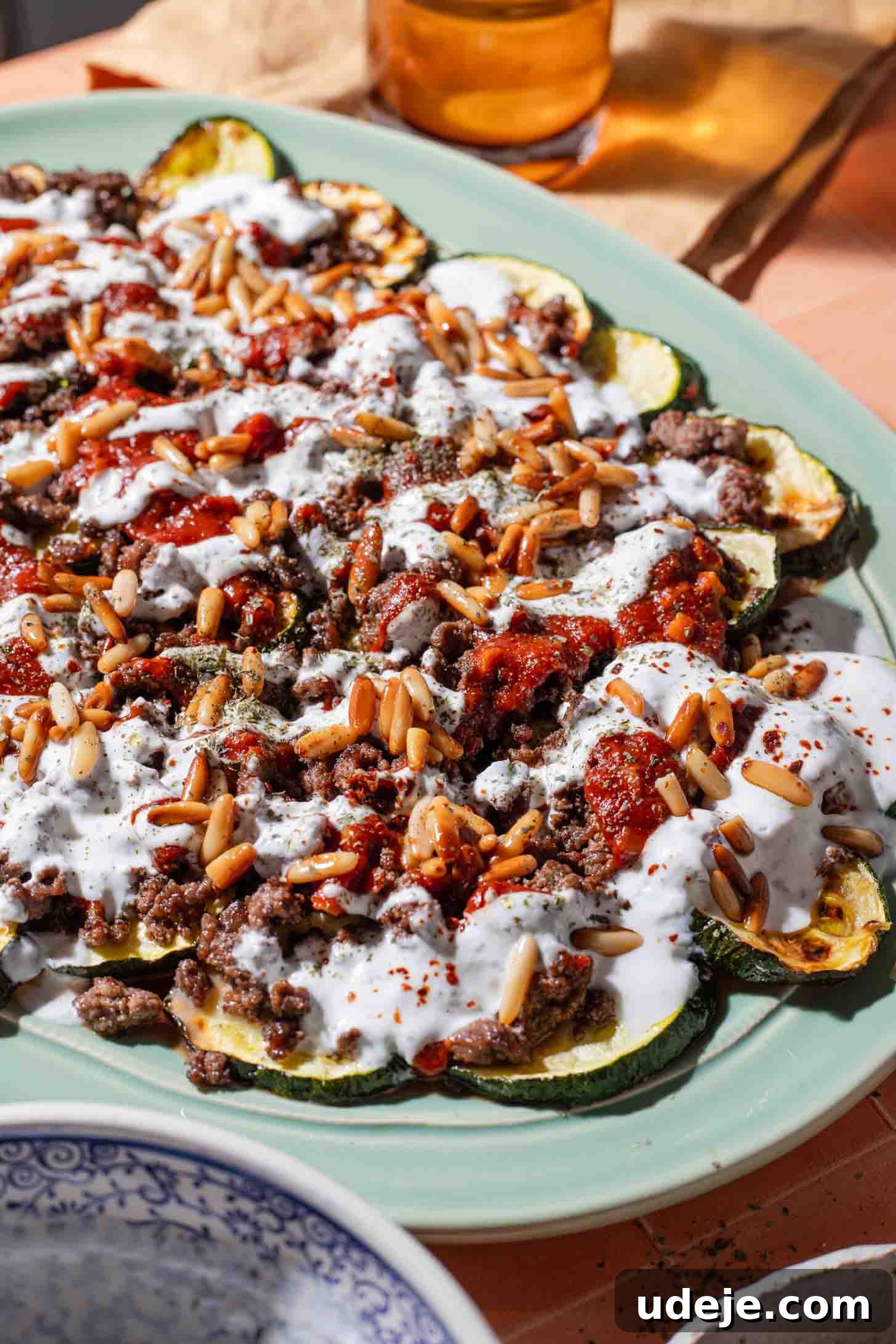 A plate of roasted zucchini rounds topped with ground meat, tomato sauce, yogurt sauce, toasted pine nuts, and herbs on a light green dish.