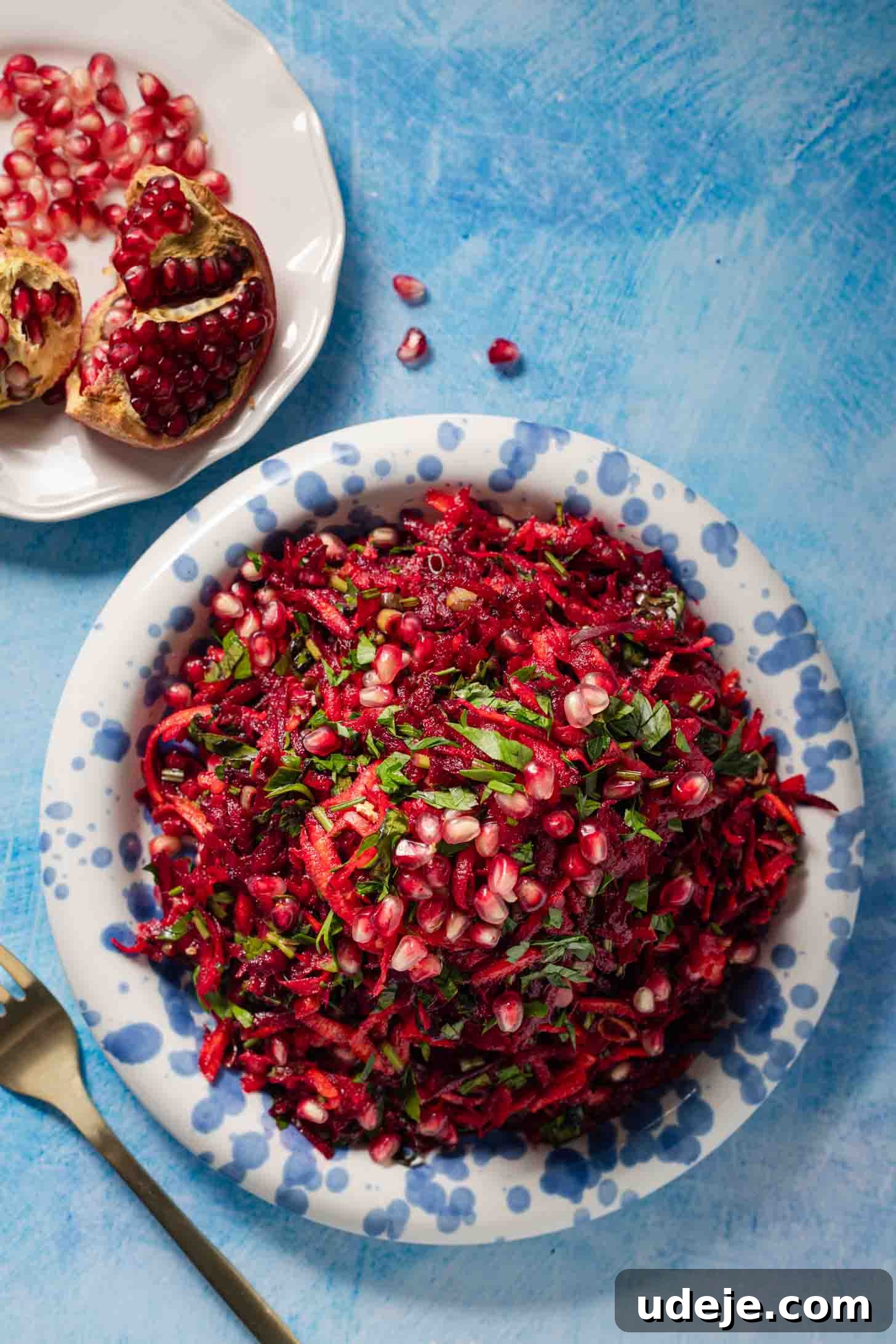 A plate of shredded beet and carrot salad topped with pomegranate seeds and herbs sits on a blue background. A fork and a halved pomegranate are beside the plate.