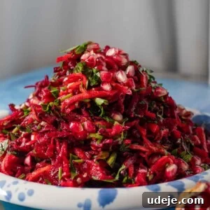 A plate piled with shredded beet salad, garnished with fresh herbs and pomegranate seeds, sits on a blue surface. A pomegranate half is visible in the foreground.