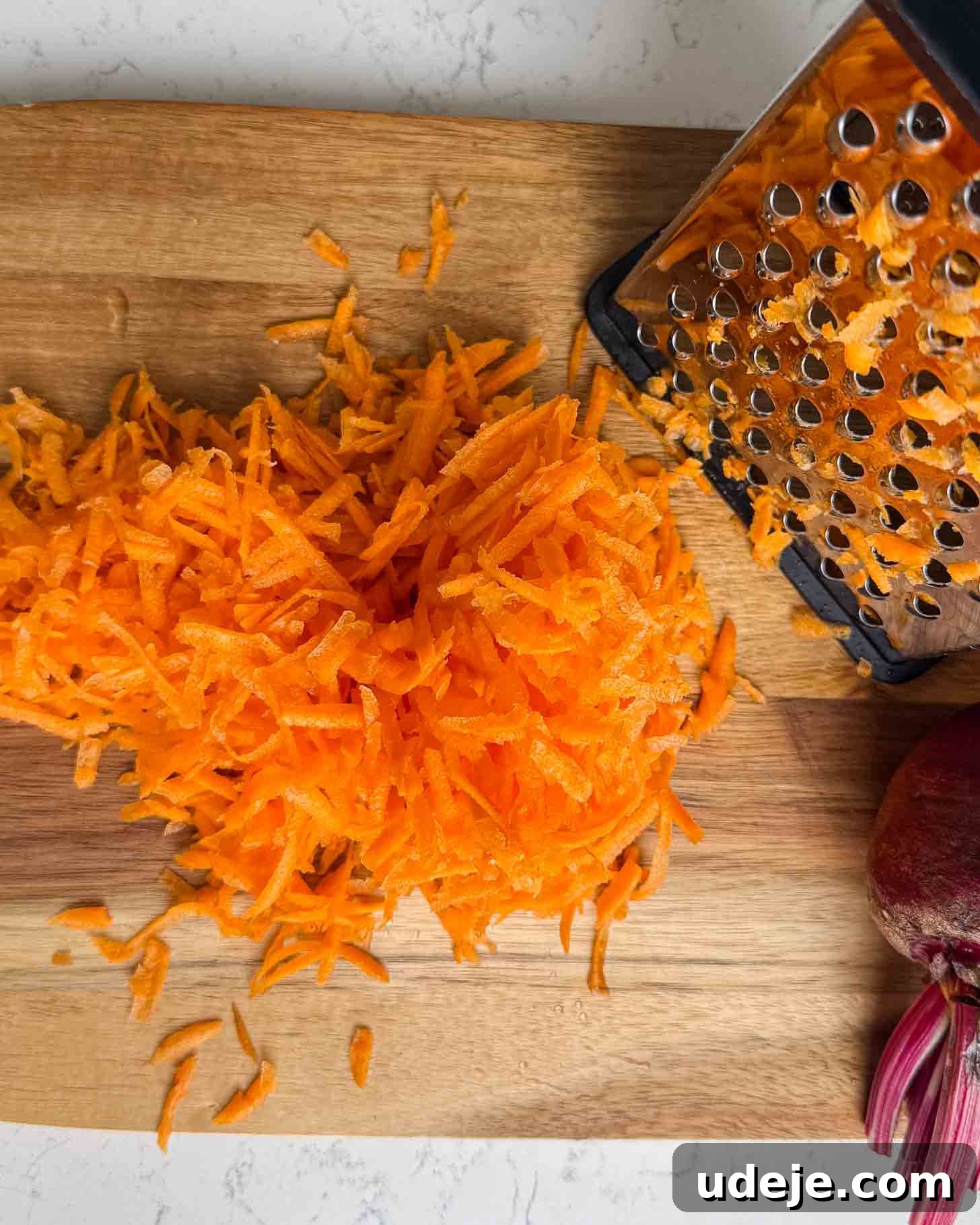 A pile of freshly grated carrots on a wooden cutting board, next to a metal box grater and part of a red onion in the corner.