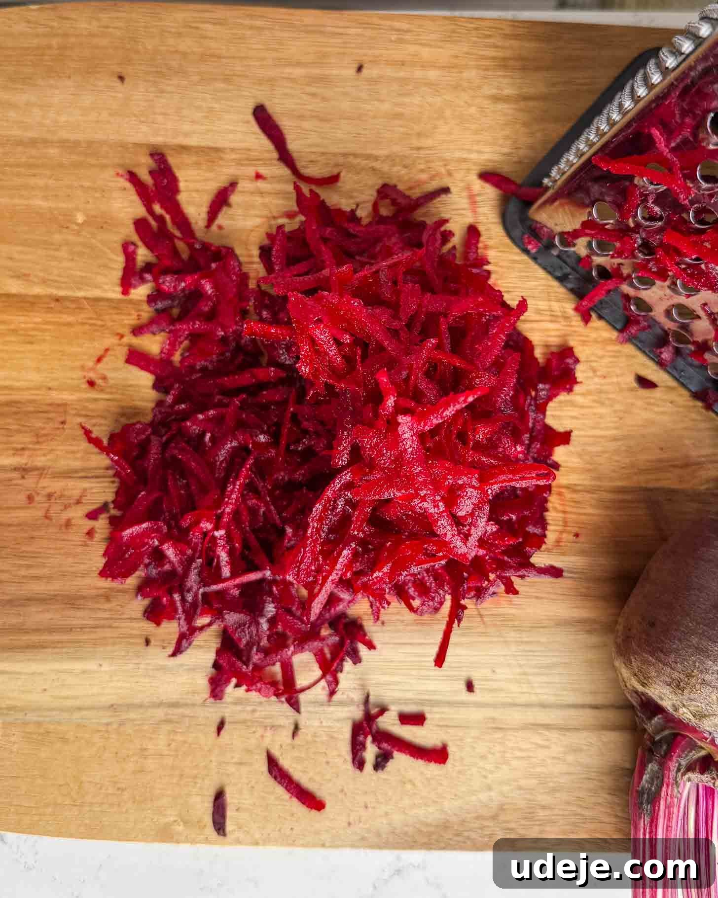 A pile of freshly grated beetroot sits on a wooden cutting board next to a metal grater and part of a whole beetroot.