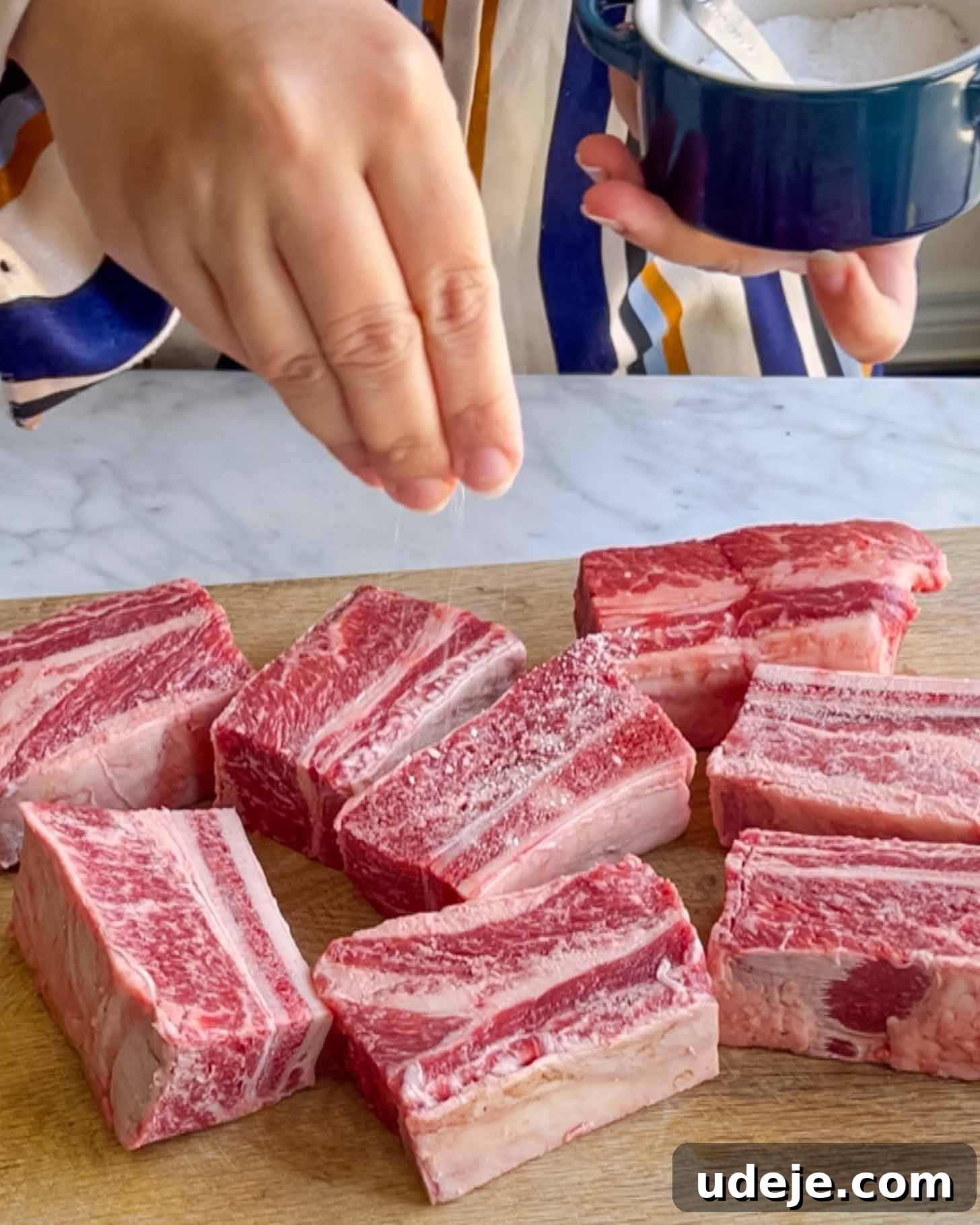 Fall-Apart Pomegranate Beef Short Ribs (No Wine) 3 A person sprinkles salt onto several pieces of raw beef short ribs arranged on a wooden cutting board.
