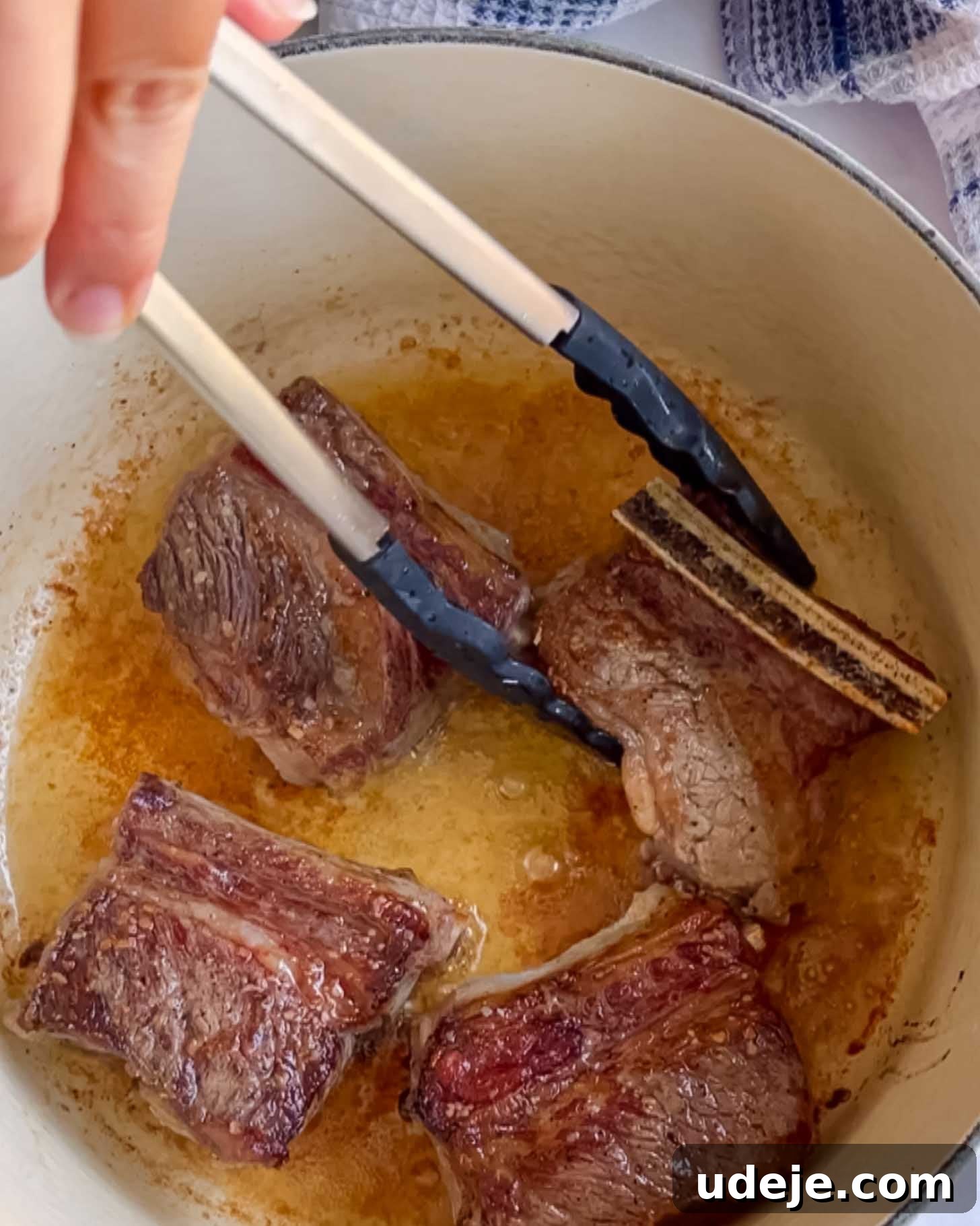 Fall-Apart Pomegranate Beef Short Ribs (No Wine) 4 A hand uses tongs to sear short ribs in a pot with oil. The meat is browning on all sides, creating a crisp exterior.
