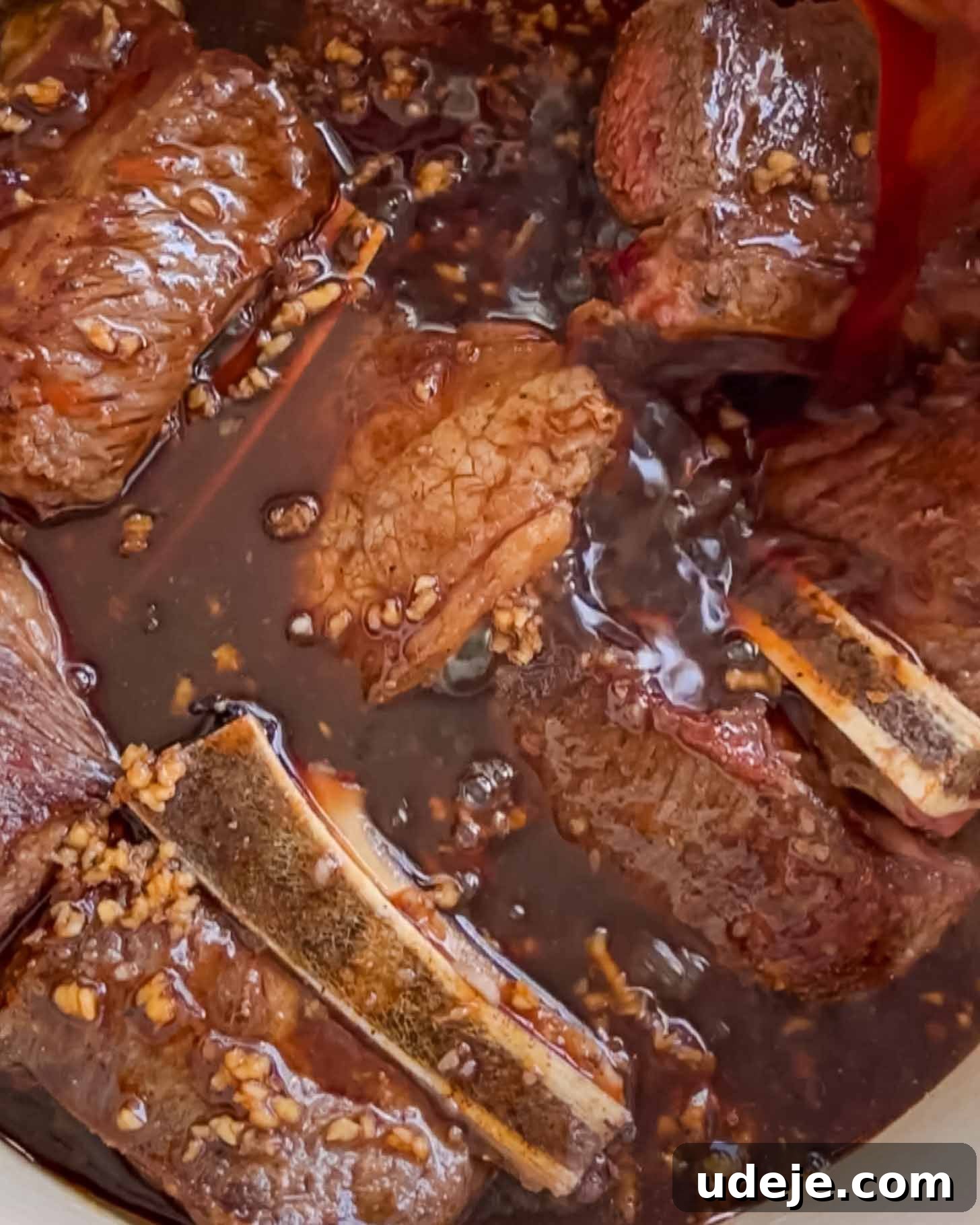 Fall-Apart Pomegranate Beef Short Ribs (No Wine) 5 Chunks of beef short ribs with bones simmer in a dark, rich sauce with visible minced garlic, creating a hearty, savory dish.