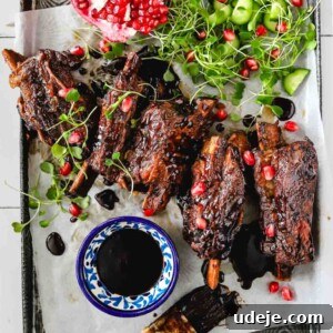 Fall-Apart Pomegranate Beef Short Ribs (No Wine) 7 A tray with glazed ribs garnished with microgreens and pomegranate seeds, next to cucumber pieces, a halved pomegranate, a bowl of dark sauce, and a sauce brush on parchment paper.